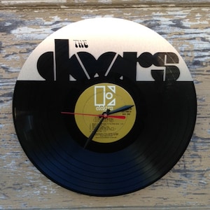 May include: A black vinyl record clock featuring The Doors album cover art. The clock face is a black vinyl record with a silver and black The Doors logo. The clock hands are red and black.