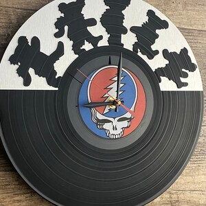 May include: A black vinyl record clock with a Grateful Dead logo and dancing bears on the top half. The clock face is white with black hands.