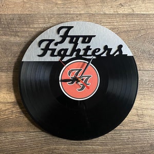 May include: A black vinyl record clock with a silver metal overlay featuring the text "Foo Fighters". The clock face is red with a stylized "FF" logo.