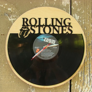 May include: A round wooden clock featuring a black vinyl record as the clock face. The words "ROLLING STONES" are cut out at the top, with the band's tongue logo. The clock hands are red and white. The record label reads "THE ROLLING STONES BLACK AND BLUE".