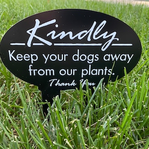May include: Black oval sign with white text that reads "Kindly Keep your dogs away from our plants. Thank You." The sign is staked into the ground in a grassy area.