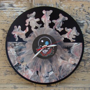 May include: A round clock made from a vinyl record. The clock face features a swirling pattern of pink, gray, and white. The center has a skull and roses design. The clock hands are white with a red second hand. Dancing bear cutouts are along the top.
