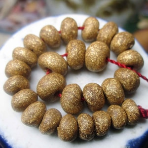 May include: A strand of gold-colored, oval-shaped beads strung on a red cord. The beads have a textured, glittery appearance. The beads are arranged in a spiral on a white and blue ceramic surface.