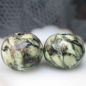 May include: Two round, marbled beads with a pale green base, black veining, and flecks of gold. The beads are displayed on a white, shell-shaped surface. These beads could be used for jewelry making.