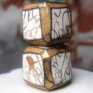 May include: Two stacked cube-shaped beads featuring a white base with black calligraphic line patterns and metallic gold painted edges. The beads have a slightly weathered texture and are positioned vertically, showcasing a hand-painted aesthetic.