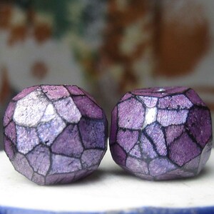 May include: Two faceted, purple beads with a geometric design. The beads have a metallic sheen and are set against a blurred background. The beads are likely for jewelry making or crafting.