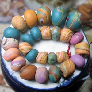 May include: A collection of colorful, handmade beads in various shapes and sizes. The beads feature a marbled effect in shades of orange, blue, pink, and brown. Strung together with a red cord, the beads are arranged in a circular pattern, showcasing their unique designs.