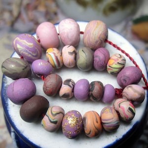 May include: A strand of handmade polymer clay beads in various shapes and colors. The beads feature swirls of purple, pink, orange, and brown. Some beads have gold leaf accents. The beads are strung on a red cord.