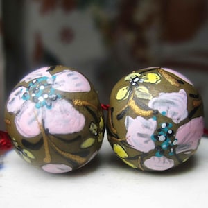 May include: Two round beads with a floral design. The beads are painted with pink, yellow, and teal flowers on a dark olive-green background. Gold accents highlight the floral details. A red string is attached.