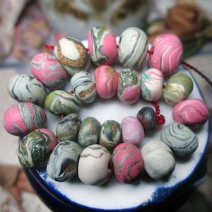 May include: A collection of handmade polymer clay beads in various sizes and shapes. The beads feature a marbled effect with colors including pink, green, white, and gold. Some beads are strung together on a red cord, showcasing their potential for jewelry making.