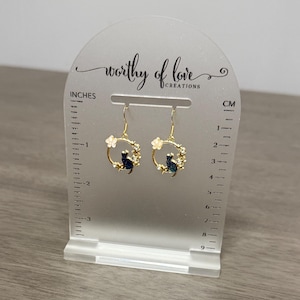 Op de afbeelding: Gouden creolen met een zwart katten- en wit bloemenontwerp. De oorbellen worden gepresenteerd op een witte acrylstandaard met een liniaal in inches en centimeters.