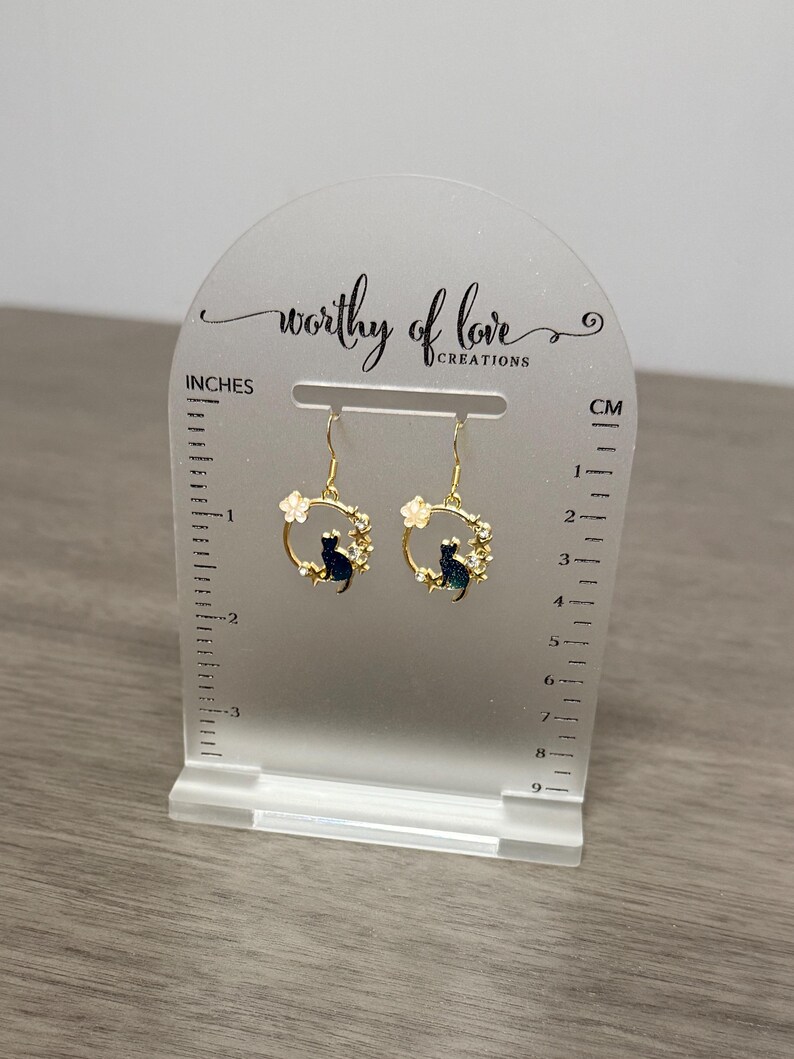 Op de afbeelding: Gouden creolen met een zwart katten- en wit bloemenontwerp. De oorbellen worden gepresenteerd op een witte acrylstandaard met een liniaal in inches en centimeters.