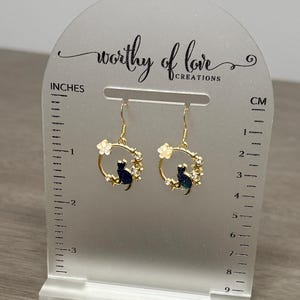 Op de afbeelding: Gouden oorhangers met een zwart katten silhouet in een cirkel van witte bloemen en strass steentjes. De oorhangers worden getoond op een witte acryl standaard met een liniaal in inches en centimeters.