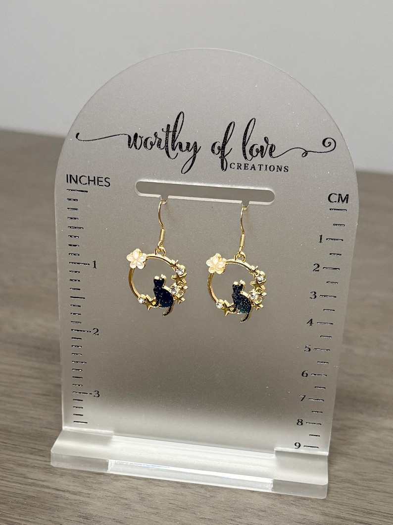 Op de afbeelding: Gouden oorhangers met een zwart katten silhouet in een cirkel van witte bloemen en strass steentjes. De oorhangers worden getoond op een witte acryl standaard met een liniaal in inches en centimeters.