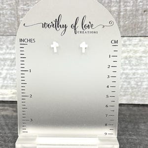 Puede incluir: Expositor de acrílico transparente con el texto "worthy of love creations" y pendientes en forma de cruz. El expositor tiene marcas de medición en pulgadas y centímetros. Los pendientes miden aproximadamente 0,6 cm.
