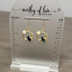 Op de afbeelding: Gouden creolen met een zwart katten silhouet en witte bloemen accenten. De oorbellen worden getoond op een witte acryl standaard met een liniaal in inches en centimeters.