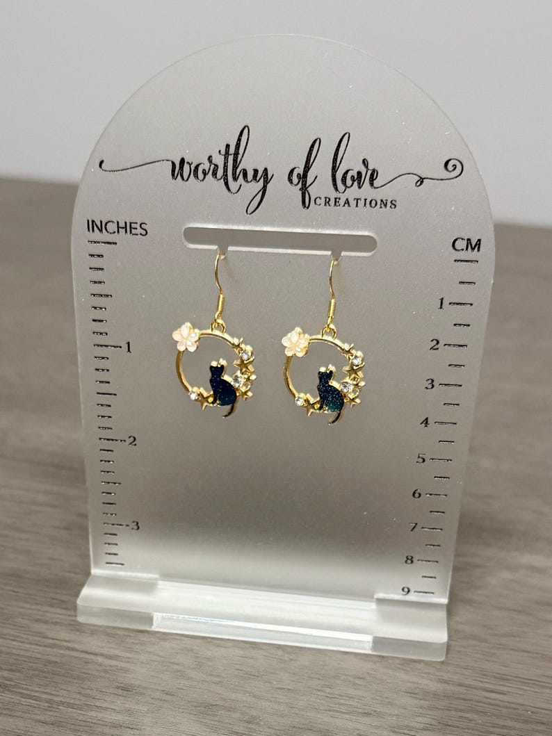 Op de afbeelding: Gouden creolen met een zwart katten silhouet en witte bloemen accenten. De oorbellen worden getoond op een witte acryl standaard met een liniaal in inches en centimeters.