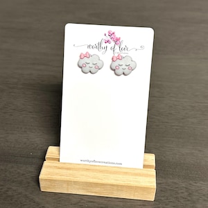 May include: A pair of white cloud-shaped earrings with pink bows and closed eyes. The earrings are displayed on a white card with the text "worthy of love creations" and the website address "worthyoflovecreations.com".