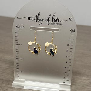Op de afbeelding: Gouden oorhangers met een zwart katten silhouet in een cirkel van witte bloemen. De oorhangers worden gepresenteerd op een witte acrylstandaard met een liniaal in inches en centimeters.