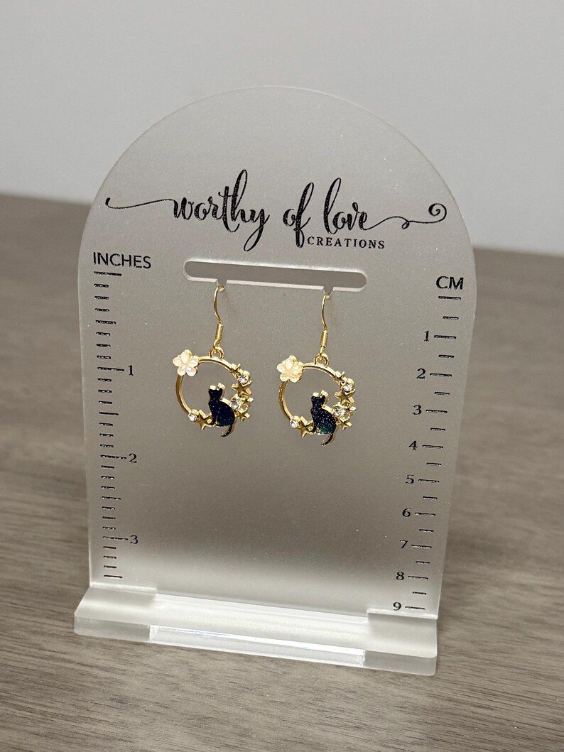 Op de afbeelding: Gouden oorhangers met een zwart katten silhouet in een cirkel van witte bloemen. De oorhangers worden gepresenteerd op een witte acrylstandaard met een liniaal in inches en centimeters.