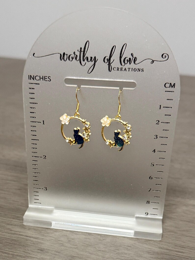 Op de afbeelding: Gouden oorringen met een kattenontwerp in zwart en groen, witte bloemen en gouden sterren. De oorringen worden getoond op een witte standaard met een liniaal in inches en centimeters.