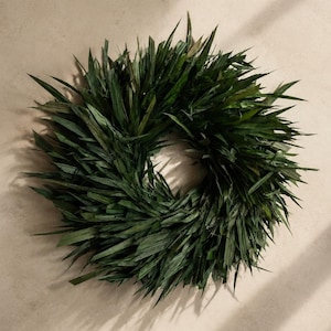 Pode incluir: Uma coroa circular construída com numerosas folhas longas e pontiagudas de cor verde-escura. A folhagem está densamente sobreposta e disposta num padrão radial, criando uma aparência cheia e texturizada com bordas afiadas na estrutura.