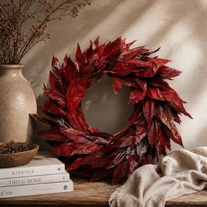 Pode incluir: Uma vibrante coroa de folhas vermelhas escuras, perfeita para decoração sazonal. A coroa é composta por numerosas folhas pontiagudas, criando uma aparência cheia e texturizada. Descansa sobre uma superfície de madeira com livros e um vaso, sugerindo um tema outonal.