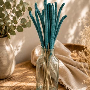 Könnte beinhalten: Eine klare Glasvase enthält einen Bund türkisfarbener Trockenblumen. Die Vase steht auf einer Holzoberfläche, mit einem neutralfarbenen Tuch und einer Keramikvase im Hintergrund. Die Anordnung wird von natürlichem Licht beleuchtet.