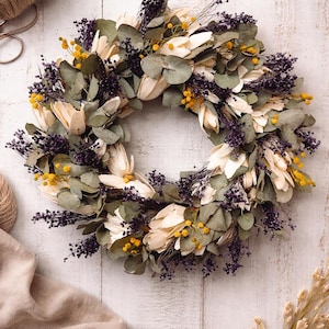 Puede incluir: Una corona circular compuesta de flores y follaje secos. La corona presenta elementos de color crema, verde, amarillo y morado. Se ven tijeras y cuerda en el fondo, lo que sugiere un tema de artesan&iacute;a o decoraci&oacute;n del hogar.