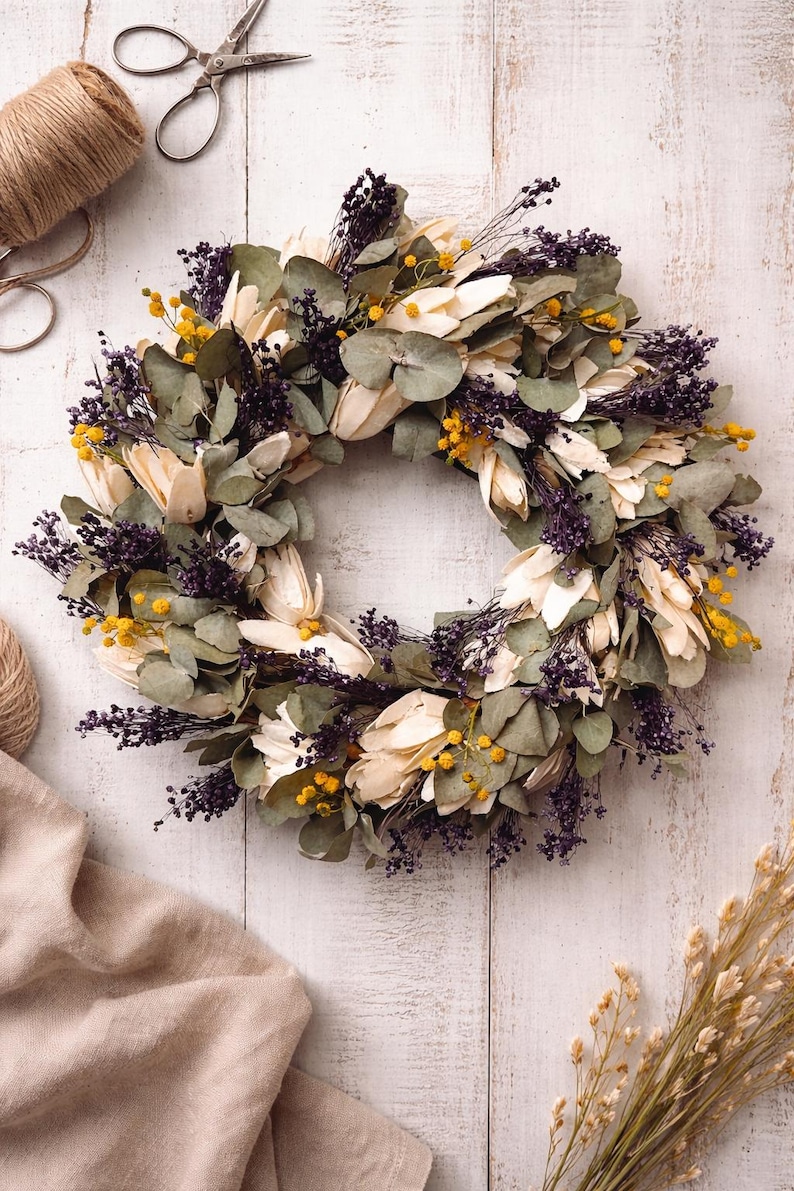 Puede incluir: Una corona circular compuesta de flores y follaje secos. La corona presenta elementos de color crema, verde, amarillo y morado. Se ven tijeras y cuerda en el fondo, lo que sugiere un tema de artesan&iacute;a o decoraci&oacute;n del hogar.