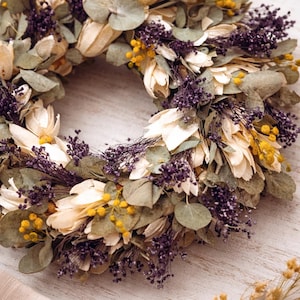 Puede incluir: Una corona floral seca de dise&ntilde;o circular. Compuesta por p&eacute;talos crema, hojas verdes, bayas moradas y acentos amarillos. La corona se exhibe sobre una superficie de madera blanca. Ideal para decoraci&oacute;n del hogar.