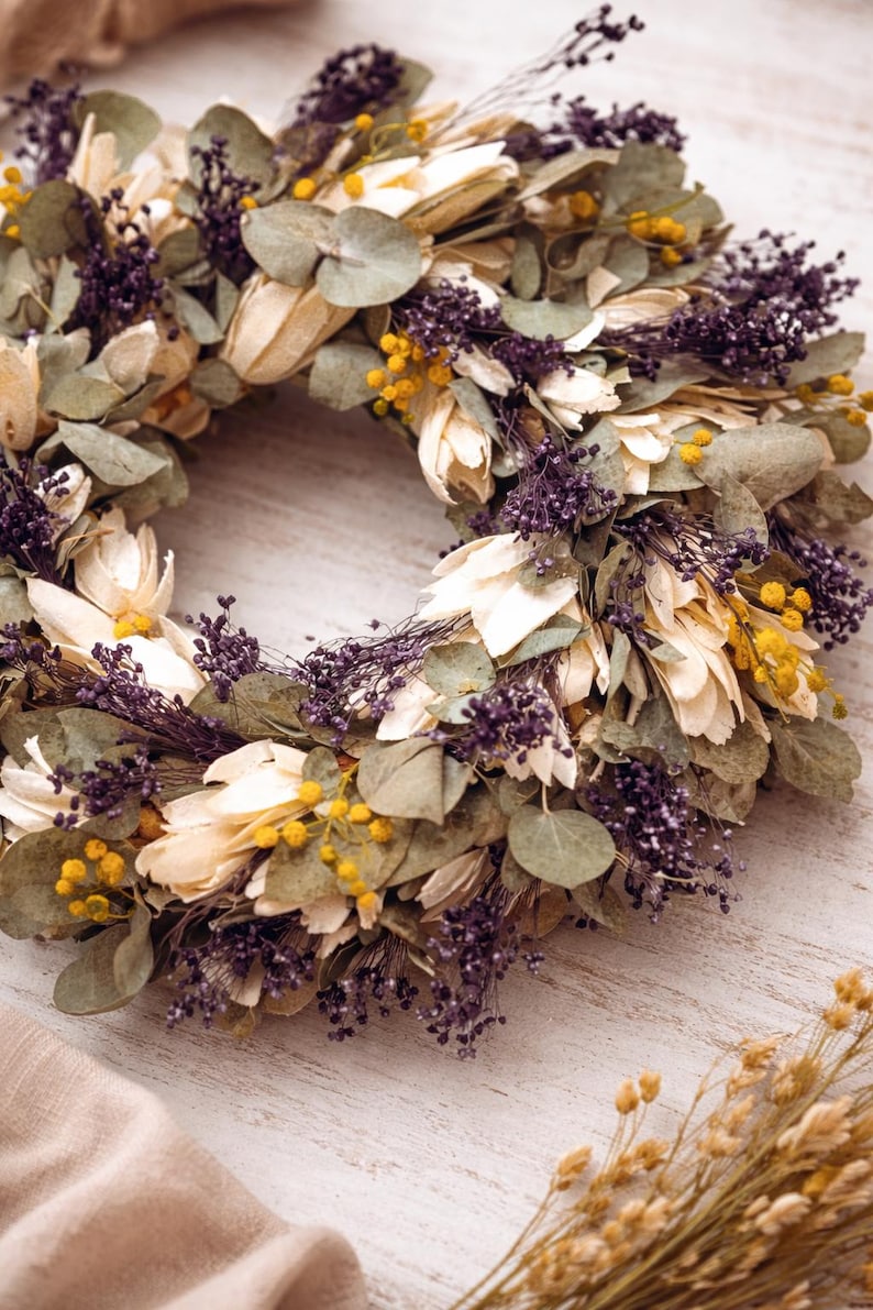Puede incluir: Una corona floral seca de dise&ntilde;o circular. Compuesta por p&eacute;talos crema, hojas verdes, bayas moradas y acentos amarillos. La corona se exhibe sobre una superficie de madera blanca. Ideal para decoraci&oacute;n del hogar.