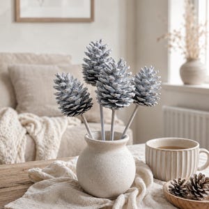 May include: A small, round, speckled beige ceramic vase containing four light blue, frosted pinecones on stems. The vase sits on a textured beige fabric cloth next to a ribbed ceramic mug and a small wooden bowl containing two additional pinecones.