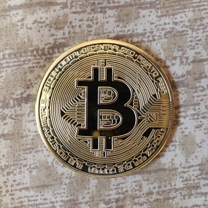 Pode incluir: Uma moeda de criptomoeda Bitcoin de cor dourada com o símbolo Bitcoin e o texto "Bitcoin: Peer to Peer Digital Decentralized System" ao redor da borda.