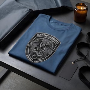 May include: A blue t-shirt featuring a graphic of a winged figure with a spear standing over a dragon inside a shield-shaped emblem. The text SLAY THE DARKNESS is printed at the top of the emblem, and Saint Michael Protect Us is at the bottom.