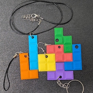 Puede incluir: Una colección de accesorios coloridos con forma de Tetris, que incluye un colgante azul en un cordón negro, un par de pendientes amarillos y un llavero con bloques morados. Otros bloques son naranjas, rojos, verdes y azules.
