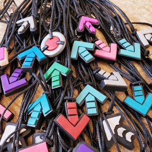 May include: A collection of colorful arrow-shaped pendants on black cords. The pendants are made of a smooth, rubber-like material and come in a variety of colors, including blue, green, red, purple, and gray.