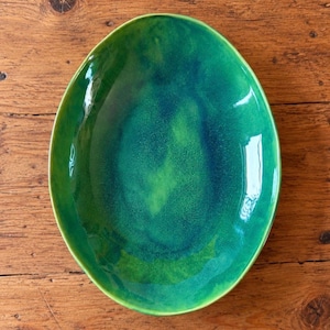 May include: An oval-shaped ceramic bowl featuring a glossy glaze with a vibrant gradient of emerald green and deep forest green tones. The surface displays a smooth, reflective finish with subtle variations in color depth and texture.