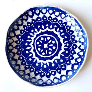 May include: A handmade ceramic bowl with a vibrant blue and white floral pattern. The bowl has a unique, irregular shape and a white rim. The intricate design features a central medallion and repeating motifs, perfect for serving or display.