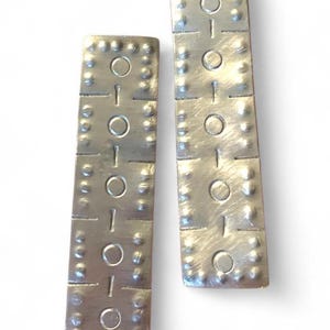 May include: Two silver-toned rectangular earrings. Each earring has a series of circles and small, raised dots. The earrings are made of a metal material and have a textured surface. The earrings are displayed against a white background.