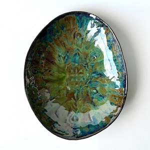 May include: An oval-shaped ceramic bowl with a glossy finish. The bowl features a blend of green, blue, and brown hues, with a dark rim. The interior has a textured, marbled appearance. The bowl is likely handmade and suitable for serving food.