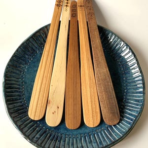 May include: Six wooden butter spreaders with decorative etched designs rest on a blue ceramic plate. The spreaders vary in color from light to dark wood tones. The plate has a textured, ridged design.