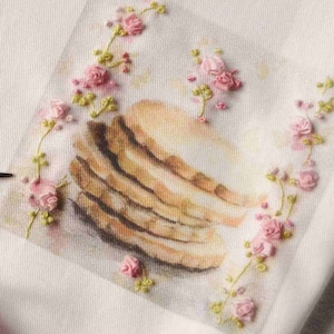 Può includere: Due pezzi di stoffa con immagini ricamate di pile di biscotti. Un pezzo di stoffa ha un nastro rosa e un paio di forbici dorate sopra.