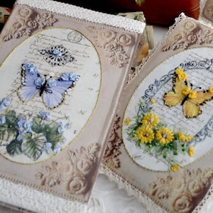 Può includere: Due copertine decorative con farfalle ricamate e motivi floreali. Le copertine sono beige con un bordo in pizzo bianco e presentano cornici ovali con testo scritto a mano.