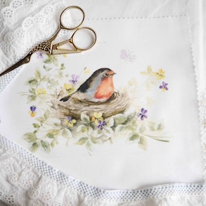 Puede incluir: Una pintura de acuarela de un petirrojo sentado en un nido rodeado de flores moradas y amarillas. La pintura está sobre una tela blanca con un borde de encaje.