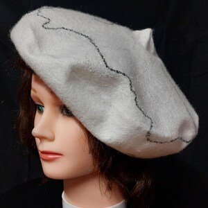 May include: A white beret with a black embroidered design. The beret is made of a soft, fuzzy material.