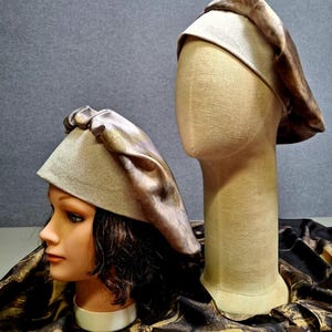 May include: Two beige berets with a brown satin-like overlay are displayed on a mannequin head and a mannequin bust. The berets have a soft, draped appearance. The background is a solid gray.