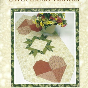 Puede incluir: Un patrón de costura para un camino de mesa llamado "Sweetheart Runner" de Thimbleberries. El patrón presenta un diseño de corazón y estrella en tonos de verde, marrón y rosa. El camino de mesa mide 20 pulgadas por 48 pulgadas.