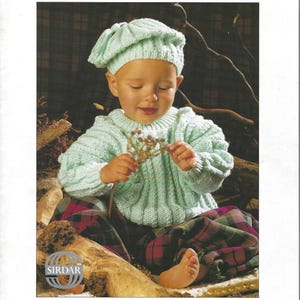 May include: A child wearing a light blue knitted hat and sweater, paired with plaid pants. The image features the text "Baby in Aran" and "Sirdar" branding, suggesting a pattern book for Aran-style baby clothing.