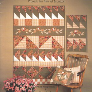 Puede incluir: Portada del libro "Lap Warmers" con proyectos para franela y algodón. Presenta un edredón grande con patrones florales y geométricos en tonos marrón, naranja y blanco. Una mecedora sostiene almohadas con diseños similares.